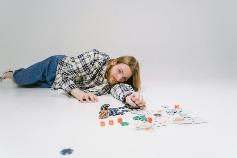 Man lying on the floor playfully surrounded by poker chips, dice, and playing cards on a white background